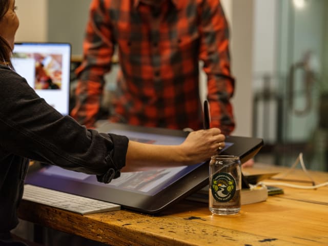 Person using a digital drawing tablet in a creative workspace, with a focus on collaboration and design, featuring a logo-embellished glass and a computer in the background.